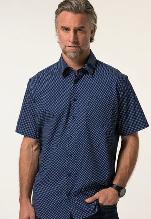 Man met grijs haar en baard, die een marineblauw kortemouwenhemd met kleine witte stipjes en een zwart polshorloge draagt, hand in zak.