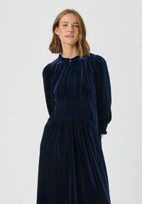 Marineblauwe fluwelen jurk met een stevige corsage, opbollende lange mouwen en een gerimpelde halslijn. Beschikt over een gerimpeld gedeelte voor een getailleerde silhoutte.