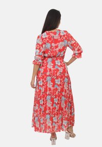 Robe maxi florale avec une base rouge, ornée de fleurs bleues et roses. Manches longues avec des poignets froncés et une taille ceinturée. Texture plissée.