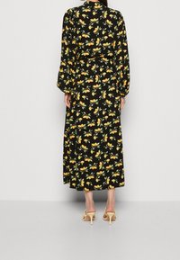 Donna che indossa un vestito lungo nero con un motivo di frutti arancioni, in piedi con la schiena rivolta verso la fotocamera, sandali con tacco beige.