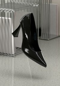 Črni patentni usnjeni salonar z ozkim nosom in koničastim peto, postavljen nad kovinsko stojalo, ki odseva svetlobo.