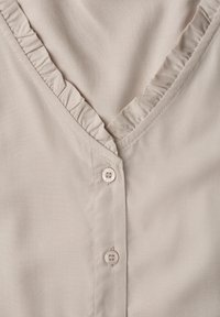 Gros plan sur un tissu beige avec un col en V à volants et deux petits boutons assortis à l'avant.