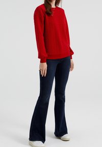 Röd texturerad tröja med ett kabelstickat mönster, med en ribbad rund halsringning och långa, puffiga ärmar. Kombineras med mörka utsvängda jeans.