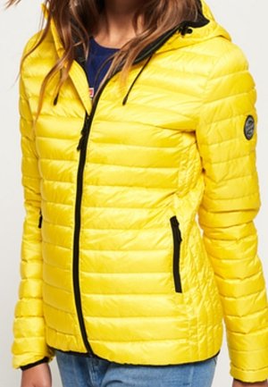 Woman wearing a bright yellow quilted jacket with black zipper and hood drawstrings, paired with blue jeans.