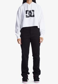 Sudadera blanca corta con el logo negro de DC, combinada con unos vaqueros negros ajustados y zapatillas negras con acentos rosas. Estilo urbano casual.