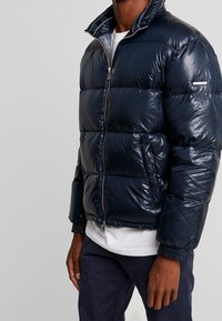 Veste matelassée marine avec une finition brillante, dotée d'une fermeture zip, d'un col montant et de poignets élastiques. Comprend des poches latérales et un design minimaliste.
