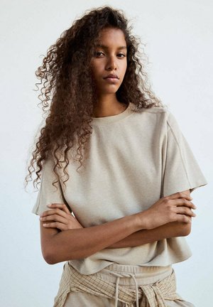 Jeune femme aux longs cheveux bouclés, vêtue d’une chemise beige à manches courtes et d’un pantalon assorti, bras croisés, expression neutre sur fond uni.