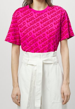 Femme portant une chemise rose vif avec un texte rouge répété "Karl Lagerfeld" et un pantalon blanc taille haute avec une ceinture en tissu nouée à la taille.