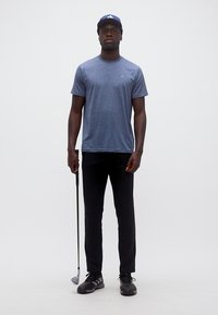 Άνδρας με μπλε καπέλο και μπλουζάκι, μαύρο παντελόνι, κρατώντας ένα μπαστούνι του γκολφ, στέκεται μπροστά σε απλό λευκό φόντο.