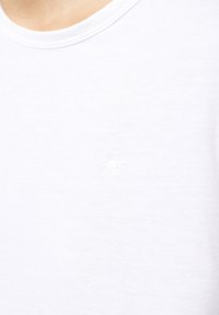Bílý bavlněný tričko s kulatým výstřihem a malým, decentním logem vyšitým bílou barvou na hrudi. Hladká textura.