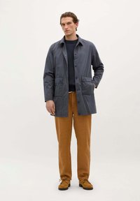Grå letvægtsfrakke med knaplukning, to forlommer, båret over en mørk struktureret sweater og tan bukser, parret med brune sko.