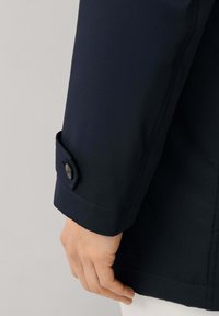 Close-up van een persoon die een donkerblauwe jas draagt met een knoopmanschette, met een ontspannen hand naast zich, tegen een effen lichte achtergrond.