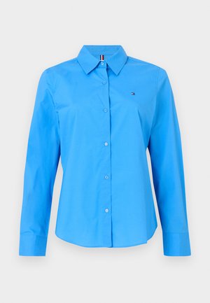 Világoskék ing, hosszú ujjakkal, galléros nyakkal és kis logóval a mellkasán. Simára szőtt anyagból készült.