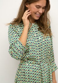 Cămașă cu model care prezintă un design în triunghiuri de culoare verde mentă, albastru marin și auriu. Confecționată dintr-un material cu o textură netedă, include butoni și o curea.