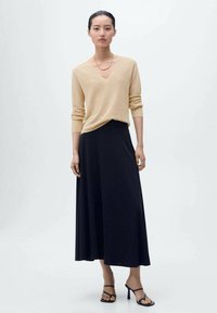 Beige gebreid truitje met een V-hals, gecombineerd met een lange zwarte rok. De outfit heeft eenvoudige lijnen en een gladde textuur, aangevuld met zwarte hakken.