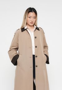 Junge Frau mit langen schwarzen und blonden Haaren, die einen beigefarbenen Trenchcoat mit dunkelbraunen Manschetten und Kragen trägt, steht mit den Händen in den Taschen.