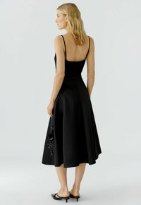 Femme aux cheveux blonds portant une robe midi sans manches noire avec des détails floraux sur la jupe, se tenant de profil devant un fond uni.