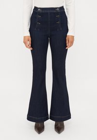 Jeans de ganga escuros de cintura alta e boca de sino, com um top justo e pernas largas, apresentando botões decorativos e costura contrastante na bainha.