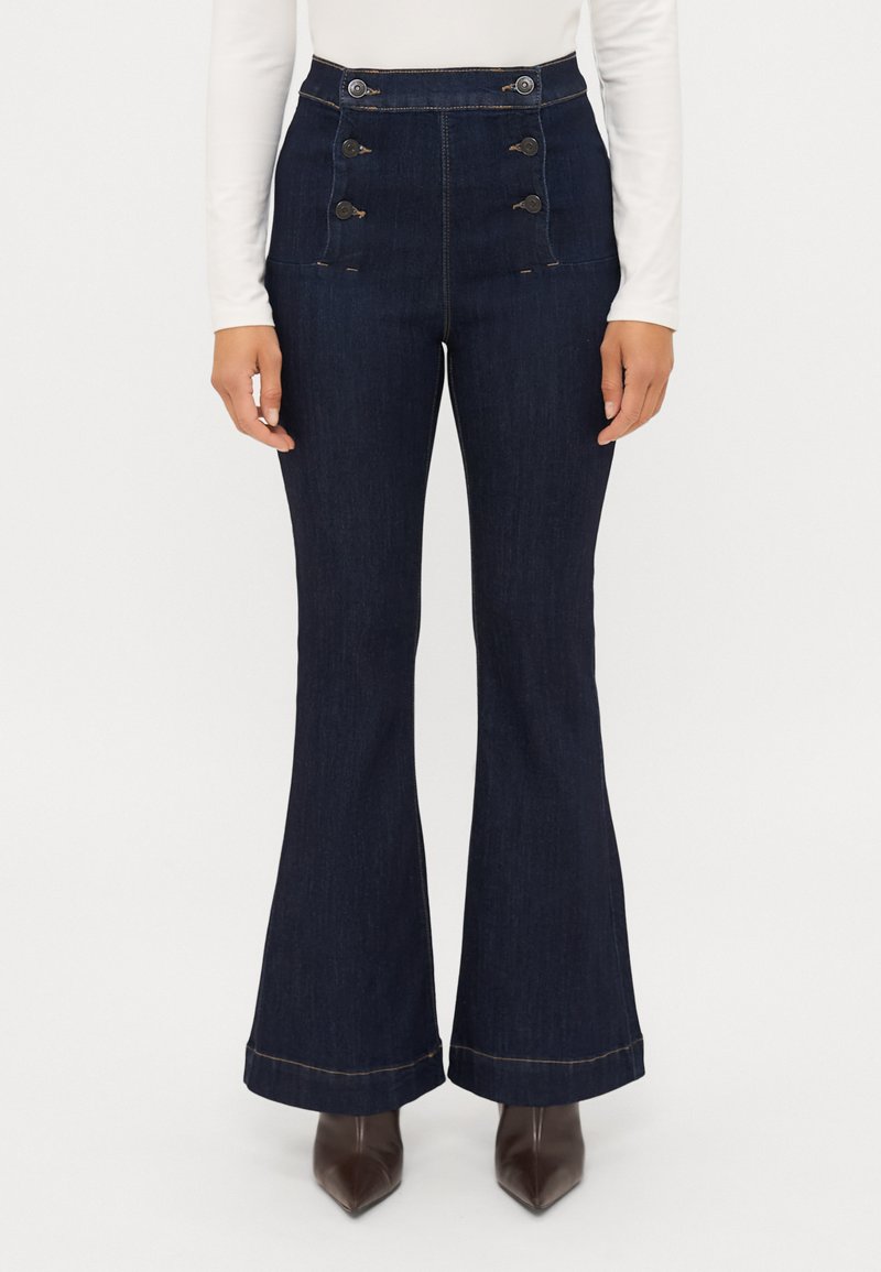 Jeans de ganga escuros de cintura alta e boca de sino, com um top justo e pernas largas, apresentando botões decorativos e costura contrastante na bainha.