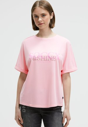 Young woman with short brown hair wearing a loose pink T-shirt with rolled sleeves and text design, paired with dark jeans with script details.
