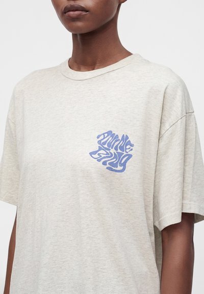 Άτομο που φοράει ένα ανοιχτό μπεζ φαρδύ t-shirt με ένα μικρό μπλε αφηρημένο γραφικό στην πάνω αριστερή πλευρά του στήθους.