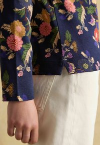 Niebieska bluzka z długim rękawem, w kwiatowy wzór, z prążkowaną teksturą i szczeliną po boku, zestawiona z jasnymi spodniami z materiału. Widoczny odcień skóry.