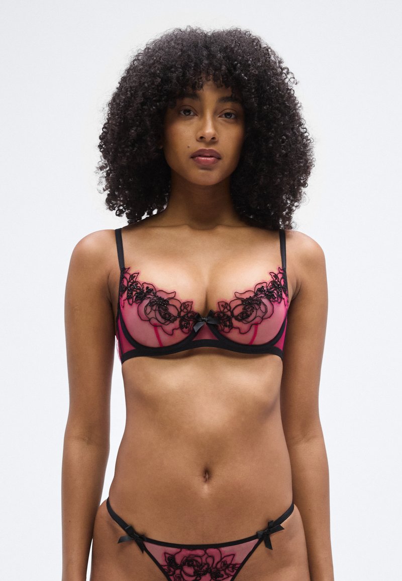 Conjunto de lencería rosa y negra con encaje, que incluye un sujetador con copas transparentes y bordado floral, acompañado de un tanga a juego con detalles de lazos.