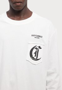 Valge pikkade varrukatega T-särk, millel on musta värvi trükitud tekst "JUST CAVALLI MILANO" ja tasku, millel on stiliseeritud must tähte "C" kujundus.