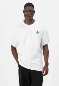 Tricou alb din bumbac cu mâneci scurte, cu un mic logo Adidas verde pe piept și un croi lejer. Asortat cu pantaloni închiși la culoare.