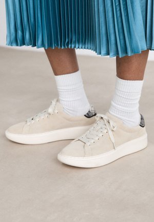 Beige suede sneakers with a white rubber sole and lace-up design. Accented with a textured black back tab. Worn with white ribbed socks.