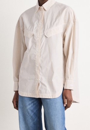 Persoon draagt een loszittend beige-wit gestreept overhemd met borstzakken, gecombineerd met een blauwe spijkerbroek, staand tegen een effen achtergrond.