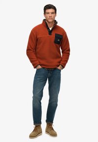 Orange fleece pullover with a black chest pocket, paired with blue jeans and tan suede boots. The collar has a contrasting inner lining.