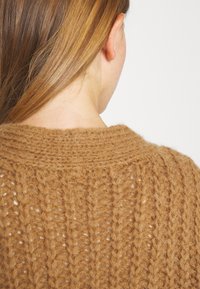 Texturiertes, braunes Strickpullover mit lockerer Passform, weitem Ausschnitt und sichtbaren Maschenmustern. Weiches, garnähnliches Material.