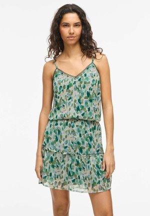 Jeune femme aux cheveux bouclés portant une robe sans manches verte et bleue à motifs avec un ourlet à volants, debout devant un fond blanc.