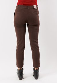 Donna che indossa pantaloni a sigaretta marroni e stivaletti neri, in piedi su uno sfondo bianco uniforme con una maglietta rossa infilata dentro.