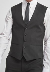 Mężczyzna w białej koszuli, czarnym kamizelce z guzikami, czarnej krawacie i czarnych spodniach eleganckich, widoczny od szyi do bioder.