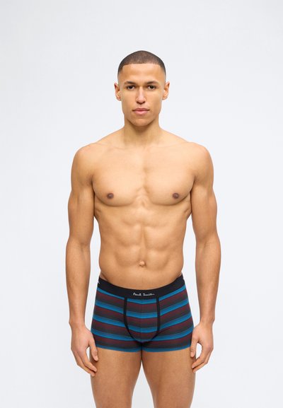 Μυώδης άνδρας με κοντά μαλλιά, φορώντας ριγωτά μαύρα, μπλε και  κόκκινα μποξεράκια, στέκεται μπροστά σε απλό λευκό φόντο.