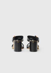 Tacchi in pelle nera con tacco largo, apertura sul retro e dettagli con cinturino. Presentano una fibbia in tonalità oro e una texture liscia.