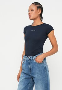 Μπλε κοντομάνικο εφαρμοστό T-shirt με στρογγυλή λαιμόκοψη, που διαθέτει ένα μικρό λευκό λογότυπο στο κέντρο. Συνδυασμένο με ανοιχτό μπλε ψηλόμεσες τζιν παντελόνες.