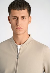 Beige zip-jacka med en mjuk textur, med rund krage och silverdragkedja, bärs över en vit skjorta. Enkel och minimal design.
