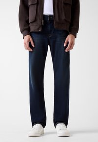 Jeans in denim blu scuro con vestibilità dritta, caratterizzati da un classico design a cinque tasche e un lieve sbiadimento. Abbinati a sneakers bianche.