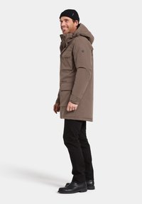 Brun parka med huva och flera fickor, tillsammans med svarta byxor och kängor. Har en strukturerad tyg och säkra stängningar.