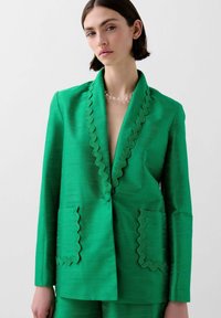 Green blazer made of textured fabric with a deep V-neck, scalloped edges, and two front pockets. Smooth, tailored fit.