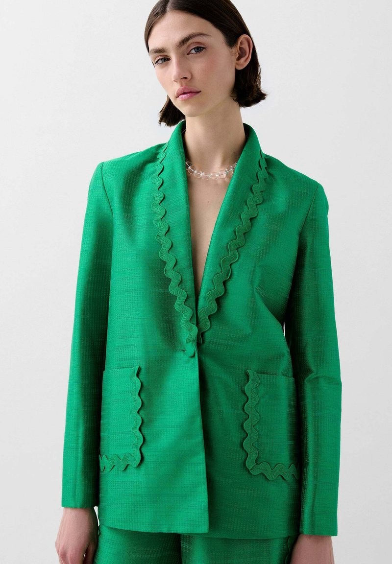 Green blazer made of textured fabric with a deep V-neck, scalloped edges, and two front pockets. Smooth, tailored fit.