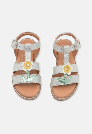 Sandales en simili cuir argentées avec trois sangles, ornées d'une appliqué de fleur jaune et de feuille verte. Semelle intérieure marron et boucles ajustables.