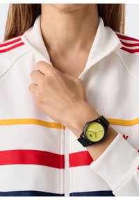 Ümar kell, millel on ere kollane nägu, must rihm ja silmapaistev must tehnika; kantud käel, millel on valge tõmblukuga jakk.