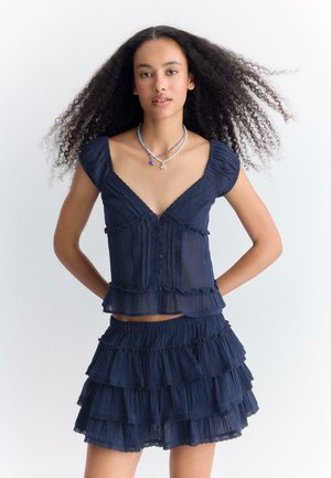 Jeune femme aux cheveux bouclés portant un haut à volants bleu marine et une jupe mini à couches assortie, debout les mains dans le dos.