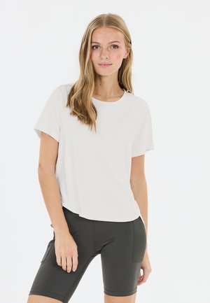 Ung kvinde med langt blondt hår, iført en enkel hvid t-shirt og sorte knælange tætsiddende shorts, stående foran en hvid baggrund.