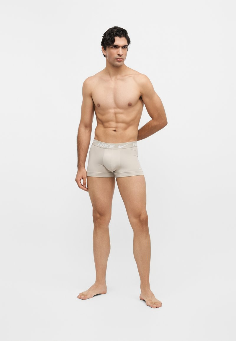 Homme en forme, debout pieds nus, portant un boxer beige Nike sur un fond uni gris clair.