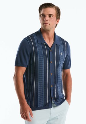 Man met een navy blauw gebreid overhemd met korte mouwen en verticale strepen, staand met één hand in zijn zak, kijkend naar links.